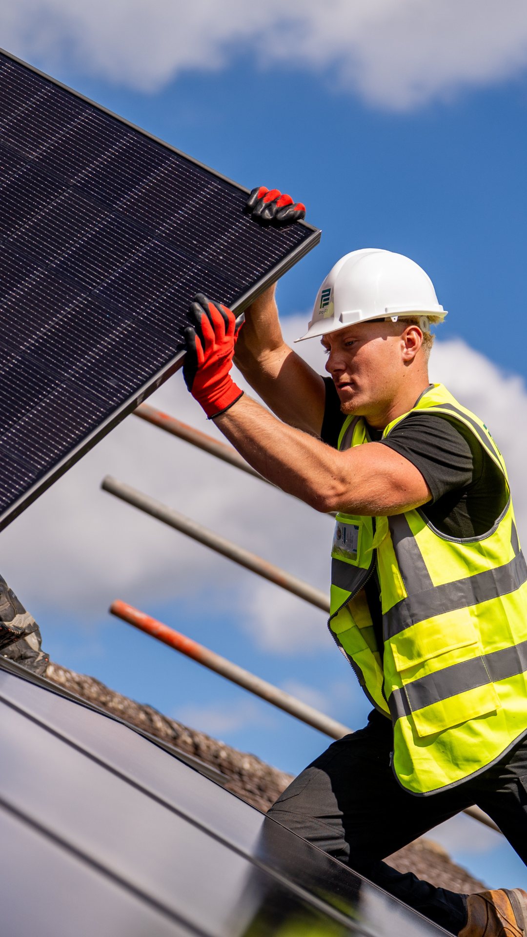 A Pro-Fit installer in a hard hat and hi-vis lifting an all-black solar panel onto a domestic roof, access platform behind him